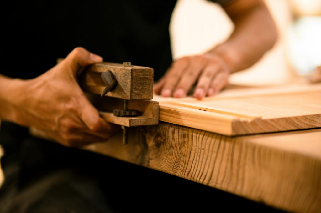 Ein erfahrener Holzhandwerker bearbeitet in einer Werkstatt Holz mit einem Handwerkzeug und demonstriert dabei handwerkliches Können und Präzision.