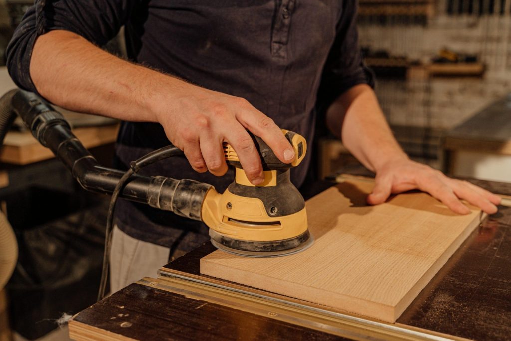 Ein Schreiner demonstriert in seiner Werkstatt sein handwerkliches Können, indem er mit einem elektrischen Schleifgerät ein Holzbrett bearbeitet.