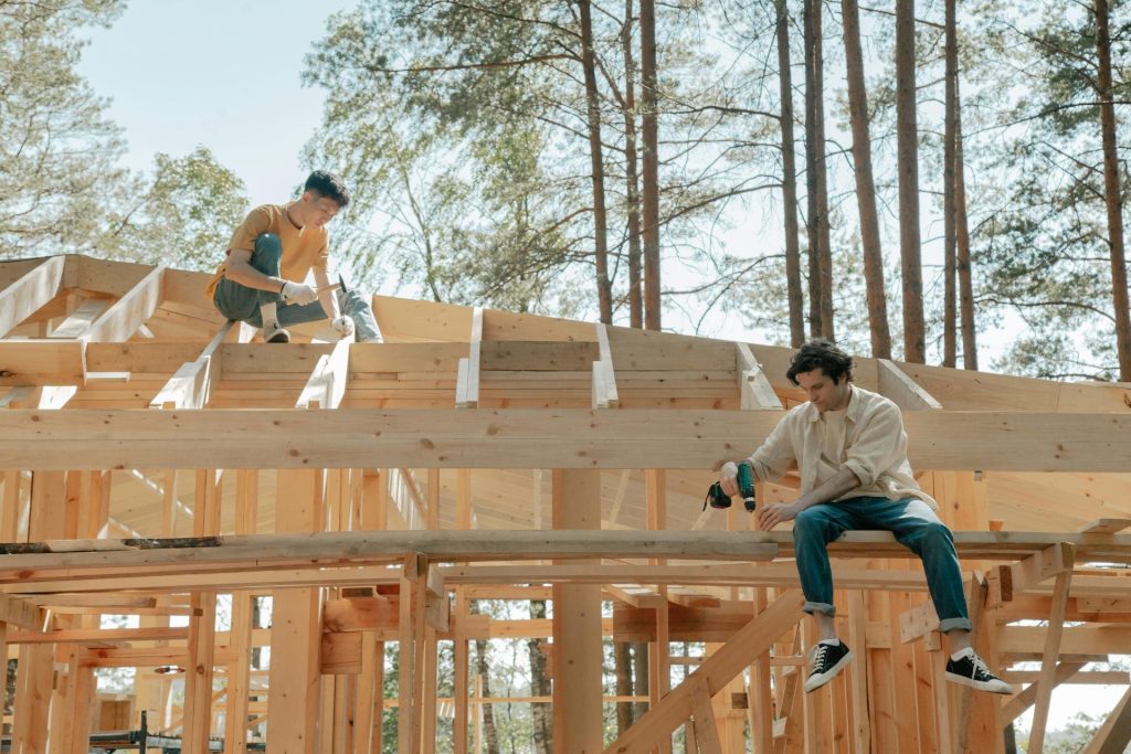 Zwei männliche Zimmerleute arbeiten bei hellem Tageslicht an einem Holzgerüst für ein Haus.